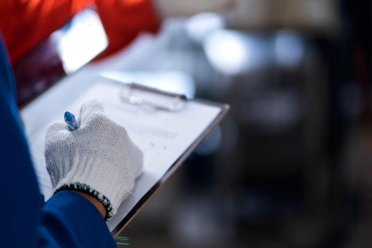 Addetta o addetto con guanto protettivo e cartellina intento a verificare documenti e mansioni in un impianto industriale metalmeccanico.